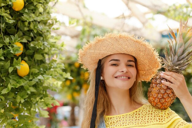 写真 草の帽子をかぶった陽気で美しい女の子が背景の夏にアナナス黄色のレモンを握っています