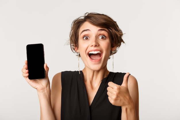 Cheerful beautiful girl showing thumbs up and demonstrating smartphone screen