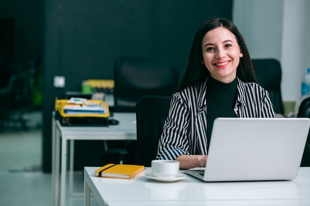 Cheerful beautiful woman in the office in front of a laptop sitting with a notebook and a cup of coffee