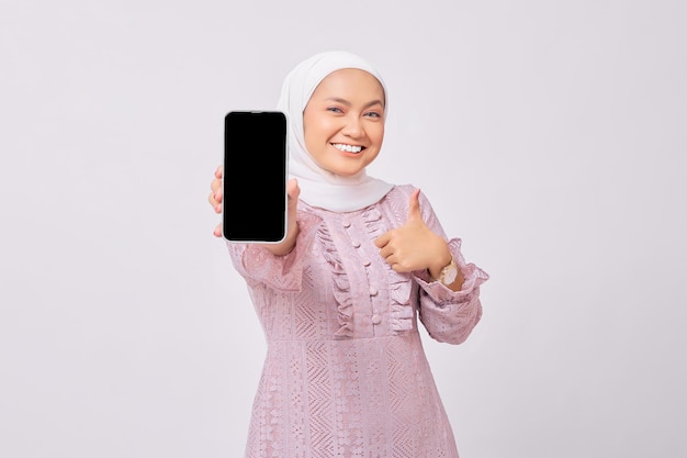 Cheerful beautiful young Asian Muslim woman wearing hijab and purple dress demonstrating smartphone with empty screen showing thumb up gesture isolated on white studio background