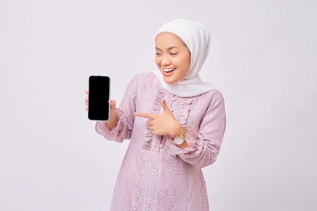 Cheerful beautiful young Asian Muslim woman wearing hijab and purple dress pointing index finger at empty smartphone screen isolated on white studio background