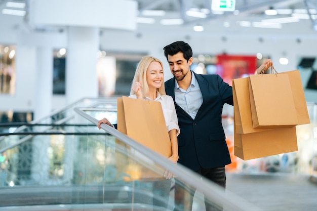 モールのぼやけた背景で購入とショッピング紙袋を持って話している陽気な美しい若いカップル