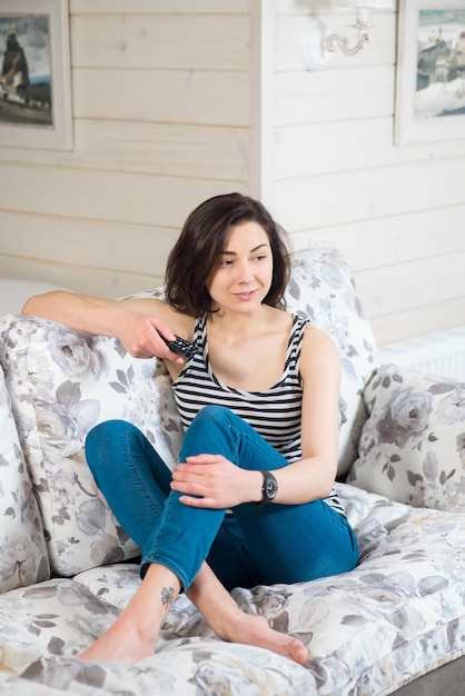 Cheerful beautiful young girl sitting on the couch and using the TV remote control at home