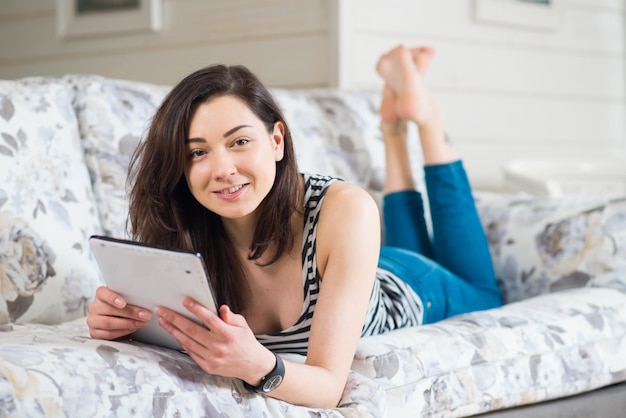 Cheerful beautiful young student girl enjoys tablet on sofa in room at home