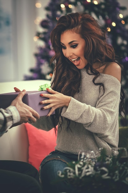 Cheerful beautiful young woman enjoying Christmas at home, opening gift and surprisingly looking what is inside.