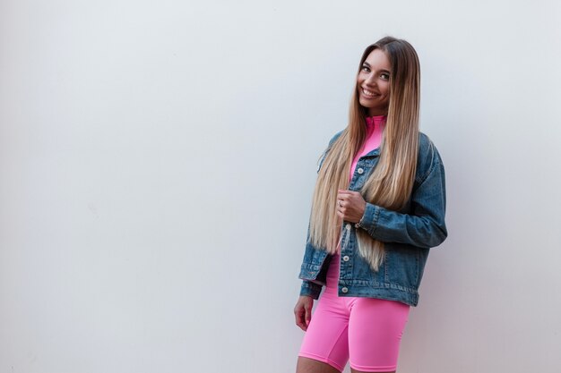 Bella giovane donna allegra in top rosa alla moda in pantaloncini rosa in una giacca di jeans con un sorriso carino si rilassa in piedi vicino a un muro vintage in strada. modello di ragazza carina felice all'aperto. stile retrò.