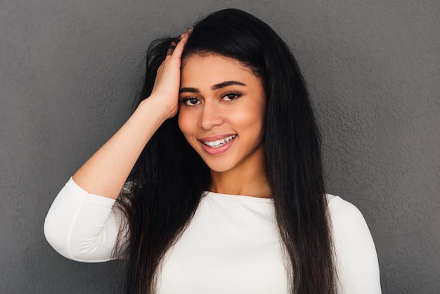 Foto bellezza allegra. attraente giovane donna africana che tiene la mano tra i capelli e guarda la telecamera con un sorriso mentre è in piedi su uno sfondo grigio