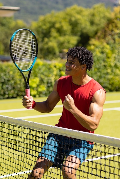 Cheerful biracial young man holding tennis racket pumping fist after winning game. Unaltered, sport, competition, hobby, nature, winner, happy, tennis court and summer concept.