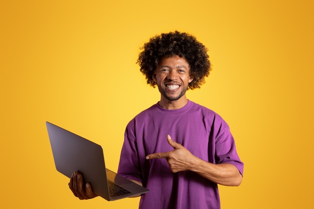 Cheerful black adult curly guy in purple tshirt points finger at laptop isolated on orange