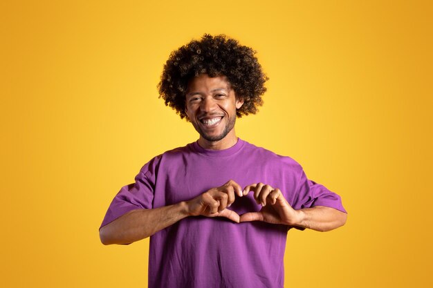 Cheerful black adult curly man in purple tshirt makes heart gesture with hands