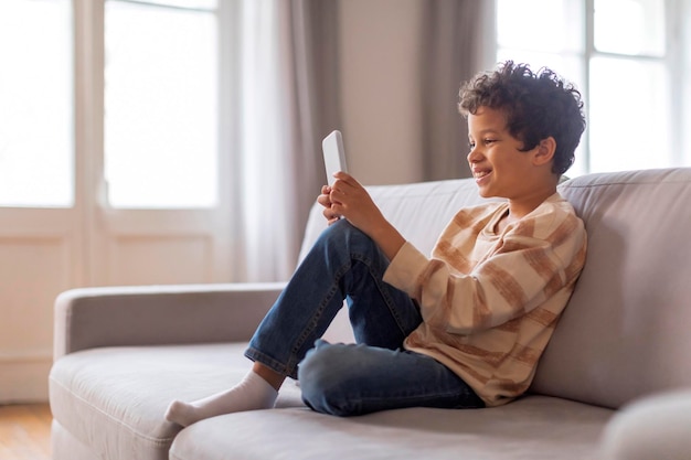 Cheerful black boy sitting relaxed on sofa deeply engaged in his smartphone