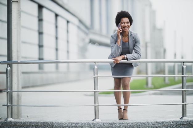 A cheerful black business woman talking on a smart phone in front a corporate building.