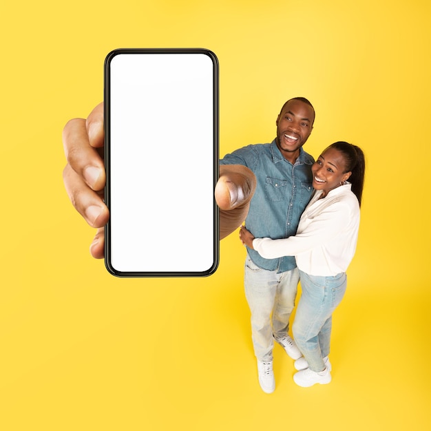 Cheerful Black Couple Showing Large Smartphone Empty Screen Yellow Background