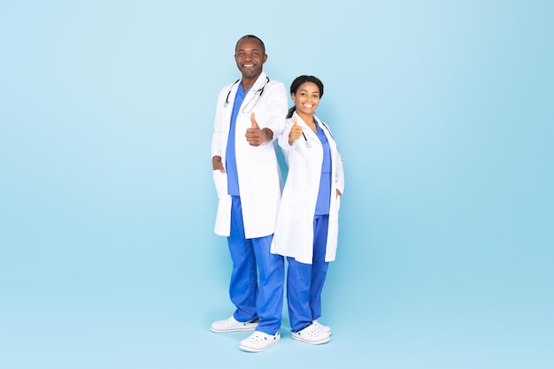 Cheerful black female and male therapists in white coats showing thumbs up blue studio background full length