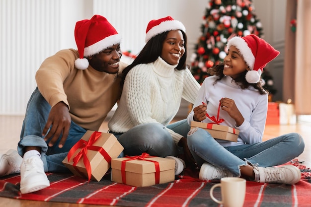Веселая черная девушка празднует Рождество с семьей, разворачивая подарочную коробку