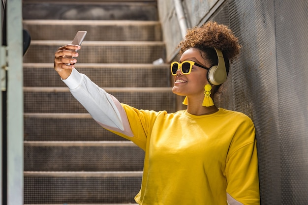 Cheerful black woman in trendy outfit taking selfie on smartphone