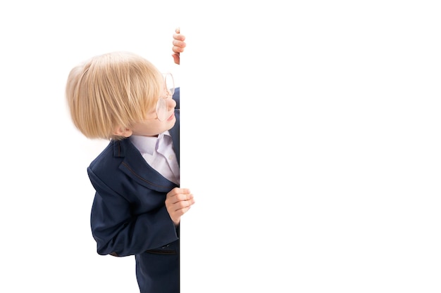 Cheerful blond boy in glasses and school uniform peeking from behind white backdrop Studio shot Copy space