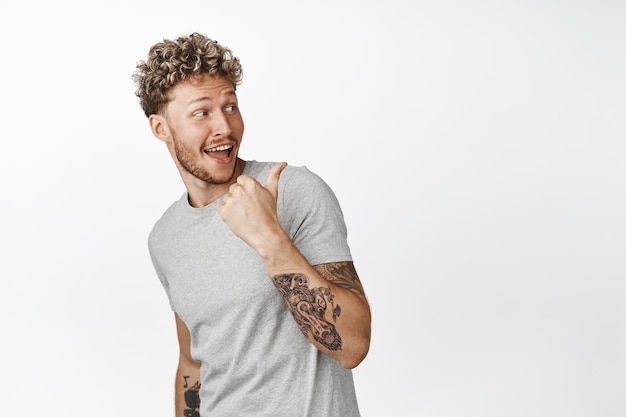 Cheerful blond curly guy turn back pointing finger behind his shoulder and smiling pleased checking out smth nice showing advertisement on background