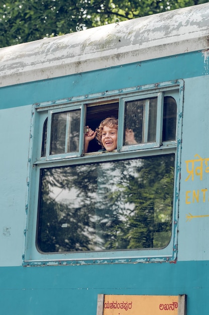 Cheerful blond kid looking out of opened window while standing in train during summer trip and looking away