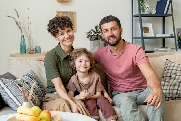 Cheerful blond little boy in casualwear sitting on couch between his happy parents during home rest in living-room