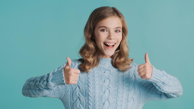 Photo cheerful blond teenage girl keeping thumbs up on camera and smiling isolated on blue background. body language concept