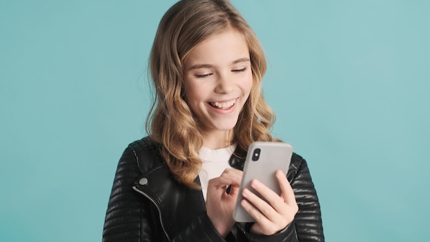 Cheerful blond teenager girl sitting in social networks looking happy isolated on blue background. Modern technology