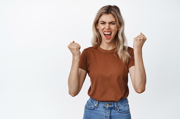 Cheerful blond woman clench fists and shouting to boost confidence encourage herself standing against white background