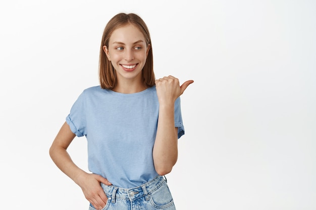 Cheerful blond woman smiling white teeth, pointing right with happy face, checking out advertisement, showing promo text, standing in t-shirt against white wall.