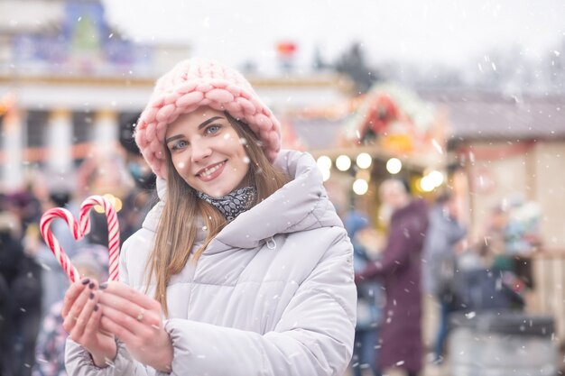 Cheerful blond woman wears pink hat and grey coat holding candy on the street Christmas fair during the snowfall. Space for text