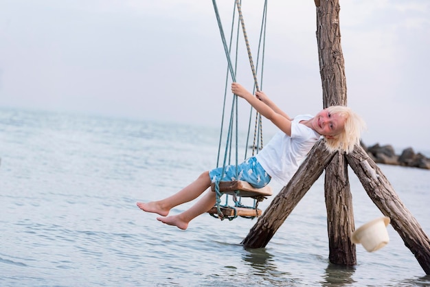 Cheerful blonde boy rides on a rope swing above sea and loses his hat Swing in the water