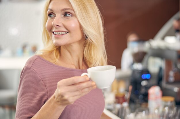 Cheerful blonde female person keeping smile on her face while looking aside at window