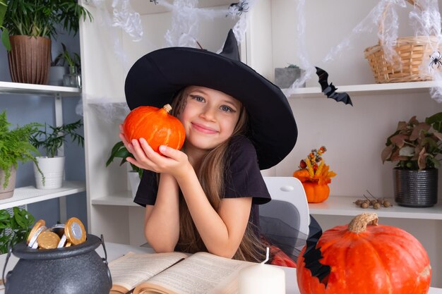 A cheerful blonde girl of 7-8 years old in a witch costume for Halloween sits at a table.