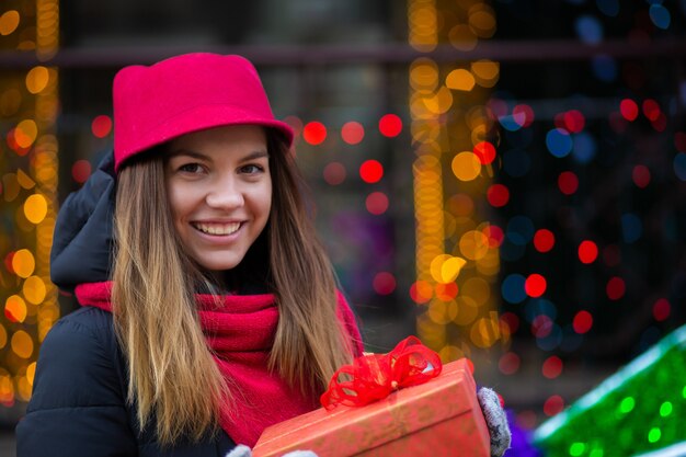 Cheerful blonde girl in red hat and knit scarf holding gift box. Space for text