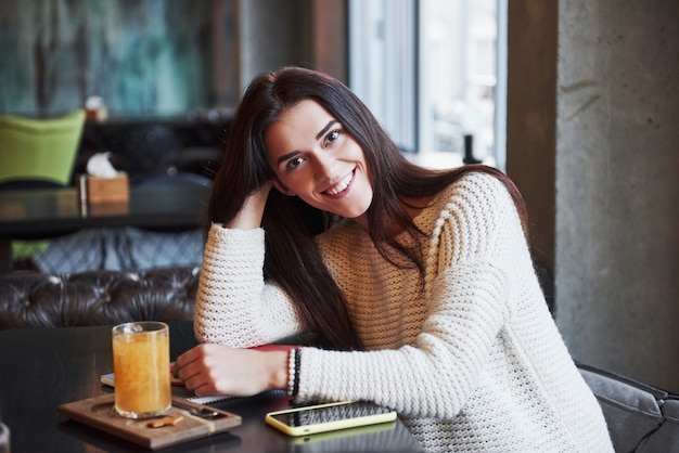 Cheerful blonde girl sit in the restaurant with yellow drink on the table and smiles