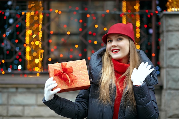 Modello biondo allegro con rossetto rosso che indossa un berretto alla moda e tiene in mano una confezione regalo con un fiocco. spazio per il testo