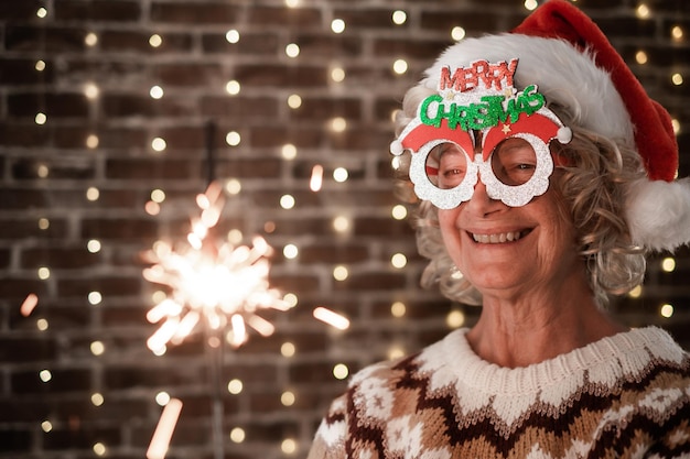 Cheerful blonde senior woman with funny glasses and Santa hat holding sparkler light elderly happy lady celebrate Christmas and new year sparkling in the night