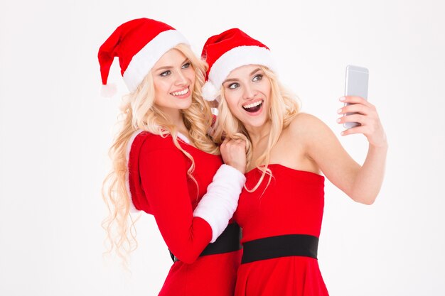 빨간 산타 클로스 옷과 모자에 밝은 금발 자매 쌍둥이 흰색 배경 위에 크리스마스 트리 근처 포즈 숨기기