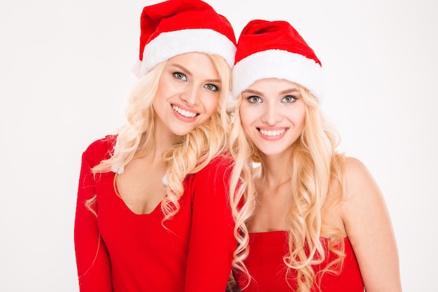 Cheerful blonde sisters twins in red santa claus clothes and hats hiding posing near Christmas tree over white background