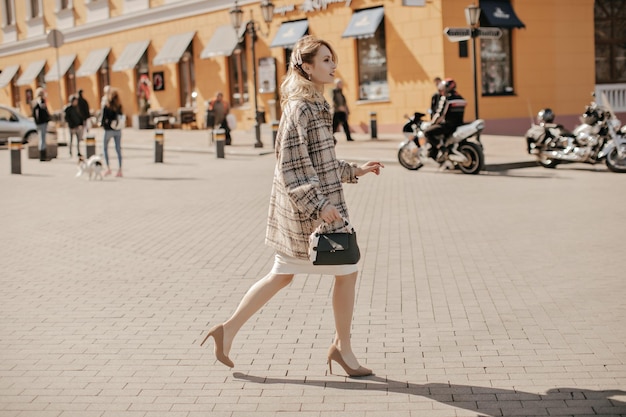 Cheerful blonde stylish lady in high heels checkered tweed coat\
and white skirt walks outside in great mood