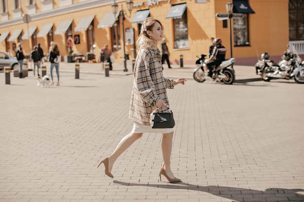 Cheerful blonde stylish lady in high heels checkered tweed coat and white skirt walks outside in great mood