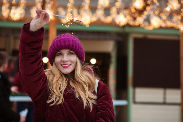 Cheerful blonde woman dressed in warm clothes, holding glowing sparklers at the Christmas fair