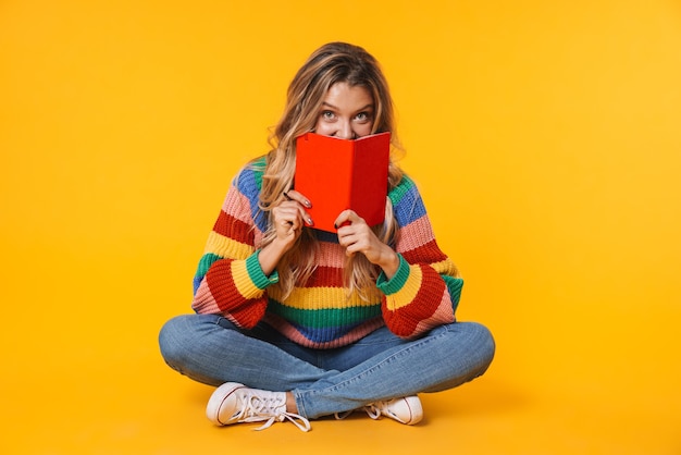 cheerful blonde woman holding diary book while sitting on floor isolated over yellow wall