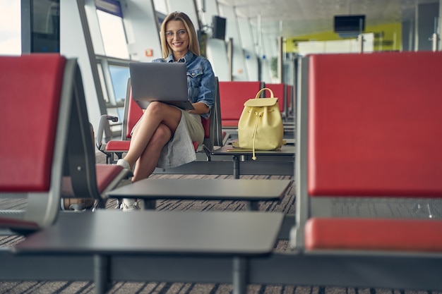 Photo cheerful blonde woman holding laptop on her knees while working on distance before flight