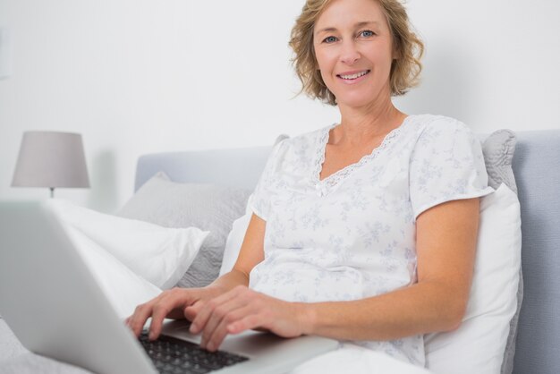 Cheerful blonde woman sitting in bed using laptop 