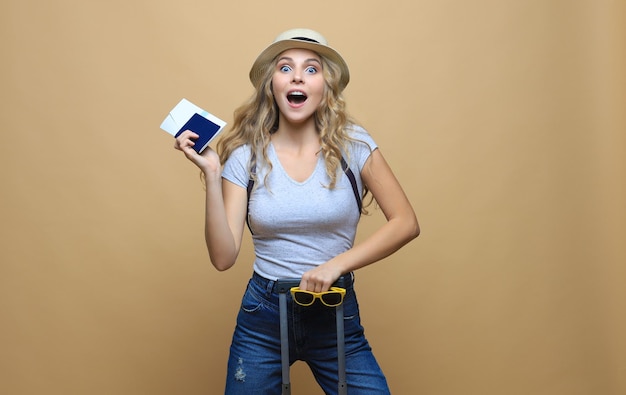 Cheerful blonde woman in summer clothes posing with baggage and holding passport with tickets over beige background.