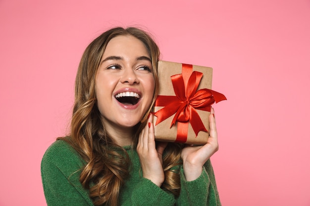 Cheerful blonde woman wearing in green sweater holding gift box and looking away over pink wall