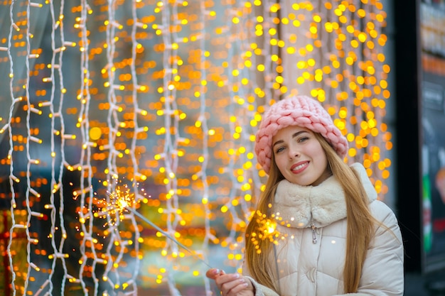 ニット帽と線香花火を保持している暖かいコートを着ている陽気なブロンドの女性