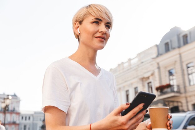 cheerful blonde woman wearing white t-shirt using cell phone and wireless earphone, while sitting in summer cafe outdoor and drinking coffee from paper cup