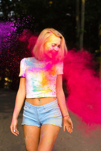 Cheerful blonde woman in white t shirt and jeans shorts playing with red Holi powder exploding around her