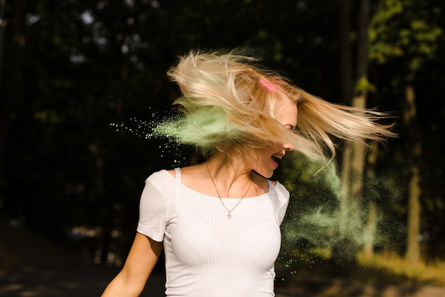 Cheerful blonde woman in white t shirt with fluttering hair posing with exploding Holi paint